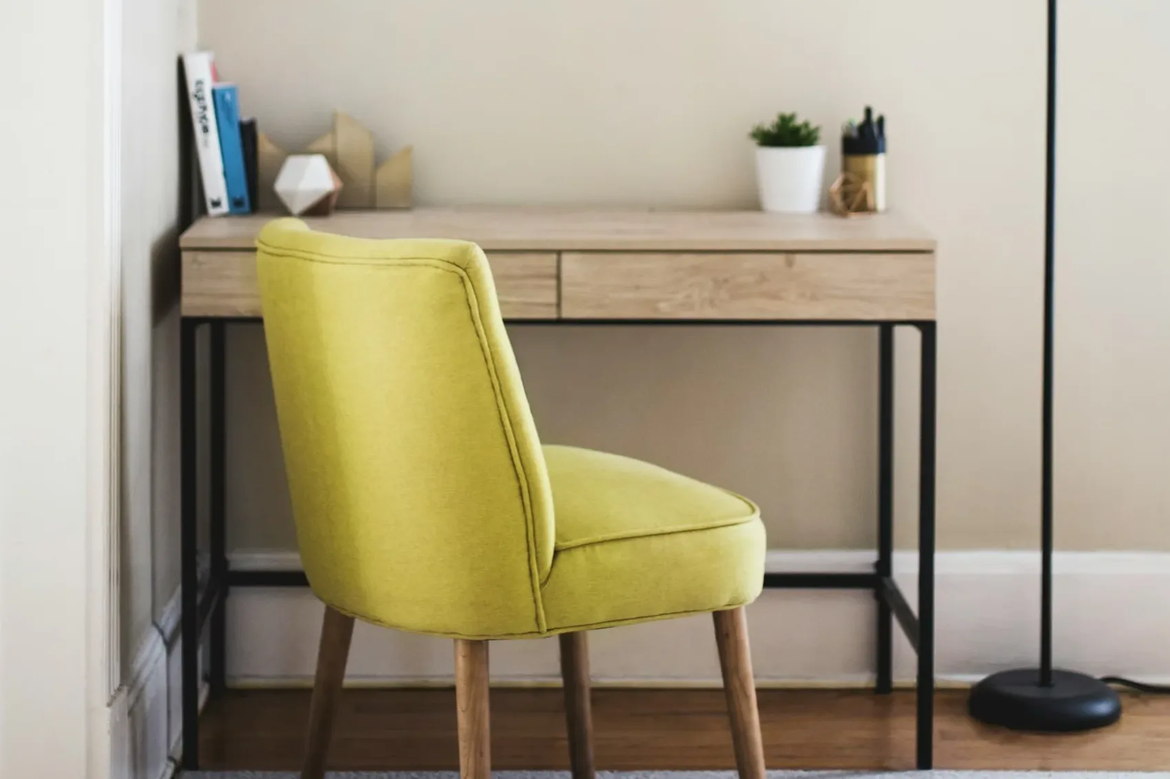 A light wood desk with a yellow chair, a lamp and a small plant against a white wall