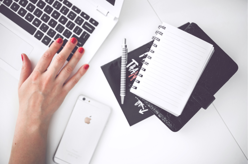 A woman typing on her laptop with a cellphone and a notebook next to the computer